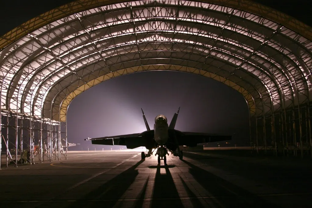 Fighter jet silhouetted inside a lit maintenance hangar at night